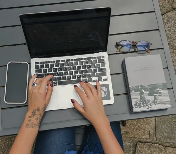 Mãos digitando em um notebook, ao lado de um livro e de óculos