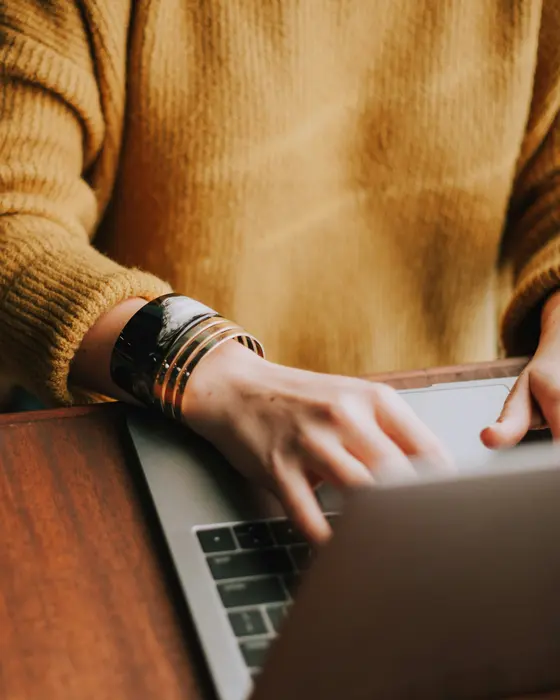 Mãos de uma pessoa de suéter amarelo digitando em um notebook sobre uma mesa de madeira
